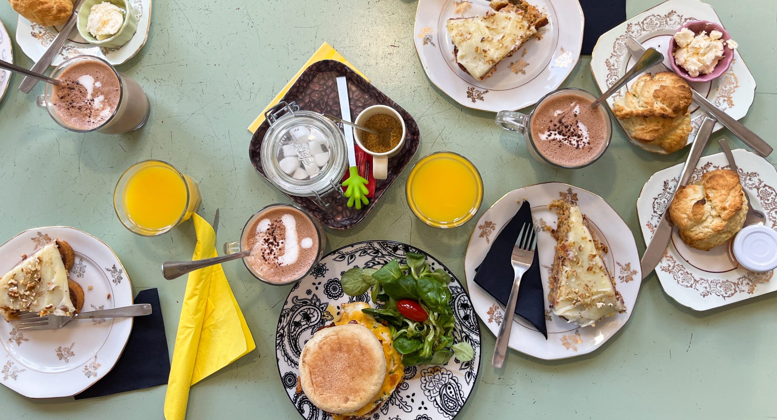 grande table avec plusieurs assiettes de desserts, de scones, ainsi que du jus d'orange et chocolat chaud